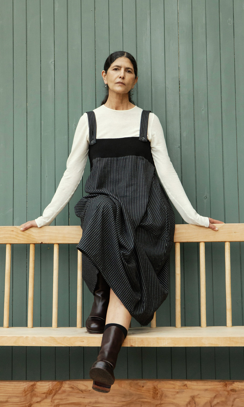 Plümo  Woman in a Pinstripe Dungaree Dress and boots sits on a wooden bench against a green paneled wall.