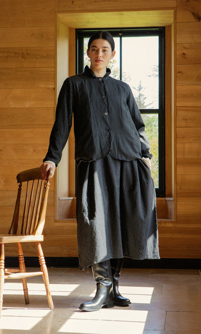 Plümo  Person in a Pinstripe Jacket stands indoors by a wooden chair, sunlight streaming through the window.