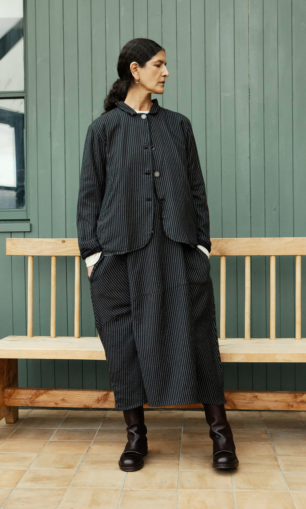 Plümo  Person in a Pinstripe Jacket stands by a wooden bench against a green paneled wall.