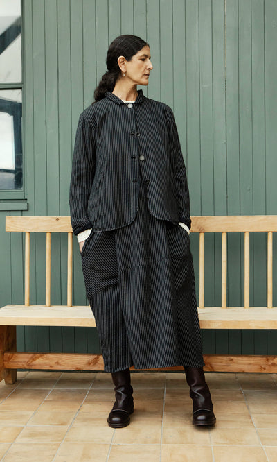 Plümo  Person in a Pinstripe Jacket stands by a wooden bench against a green paneled wall.