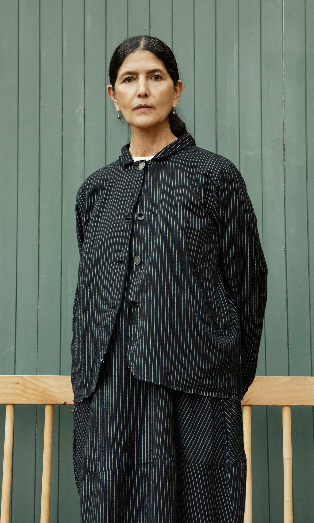 Plümo  A woman in the Pinstripe Jacket stands before a green paneled wall and a wooden bench.