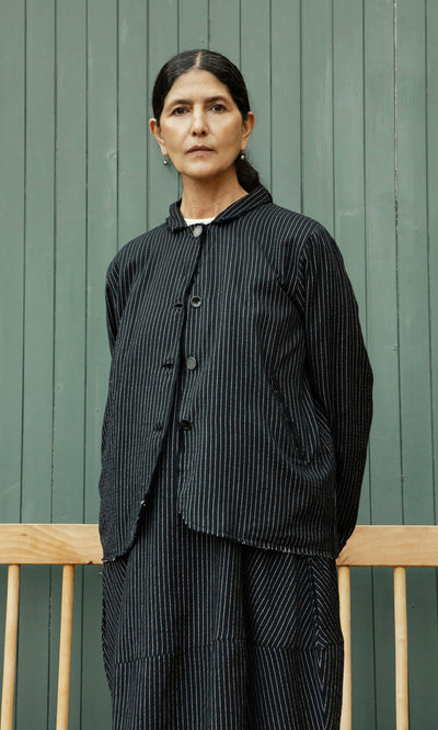 Plümo  A woman in the Pinstripe Jacket stands before a green paneled wall and a wooden bench.
