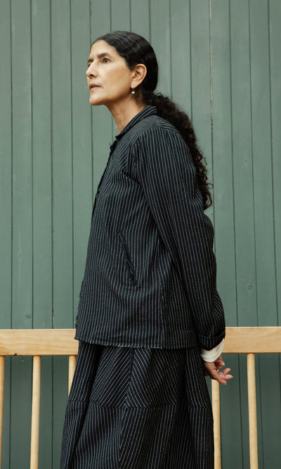 Plümo  Woman with dark hair in ponytail wears Pinstripe Jacket, standing by a green wooden wall.