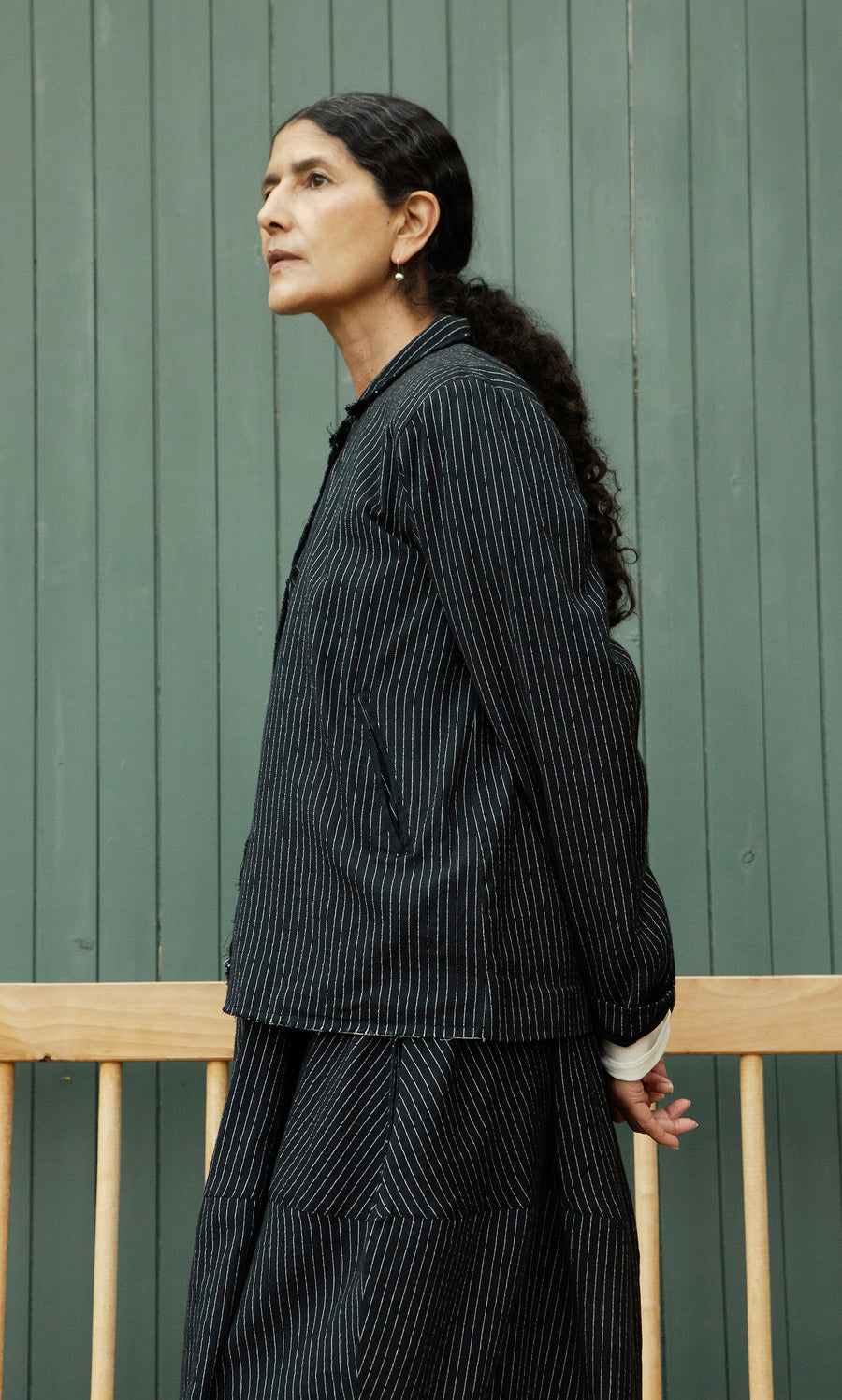 Plümo  Woman with dark hair in ponytail wears Pinstripe Jacket, standing by a green wooden wall.