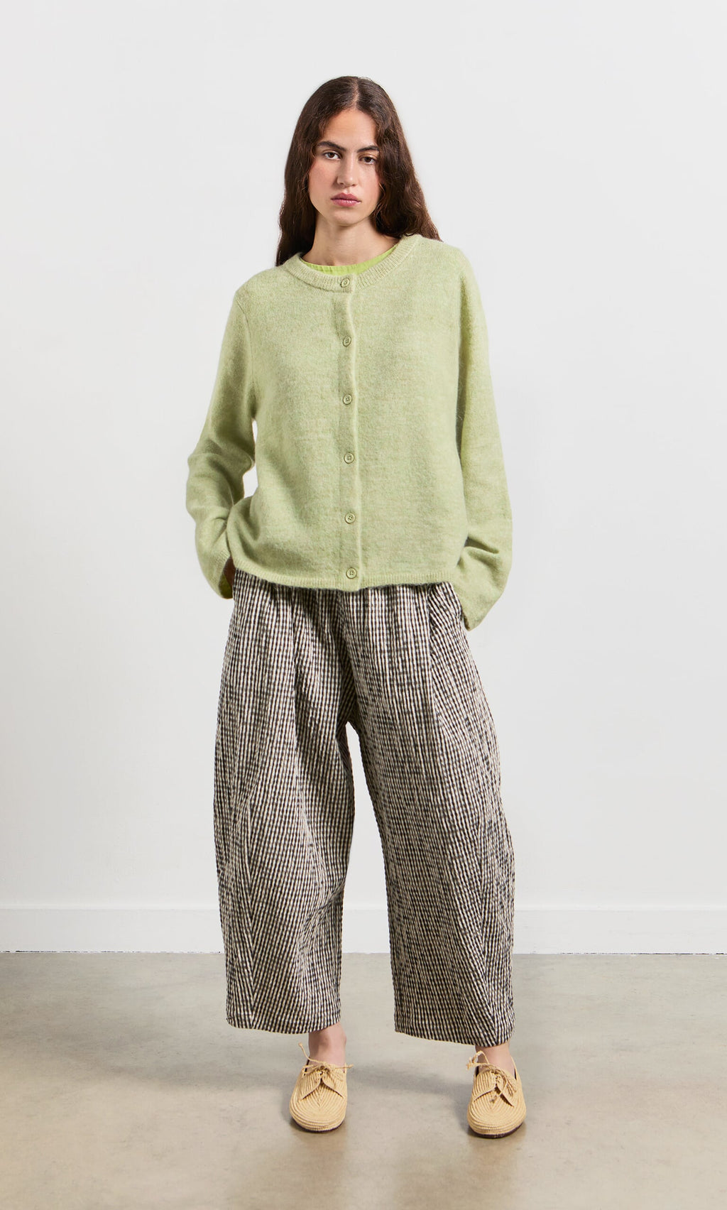 Plümo- Woman standing with hands in pockets, wearing the Pistaz Cardigan, wide plaid pants, and beige shoes.