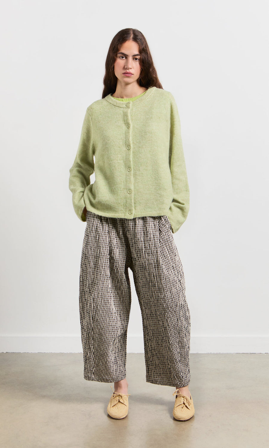 Plümo- Woman standing with hands in pockets, wearing the Pistaz Cardigan, wide plaid pants, and beige shoes.
