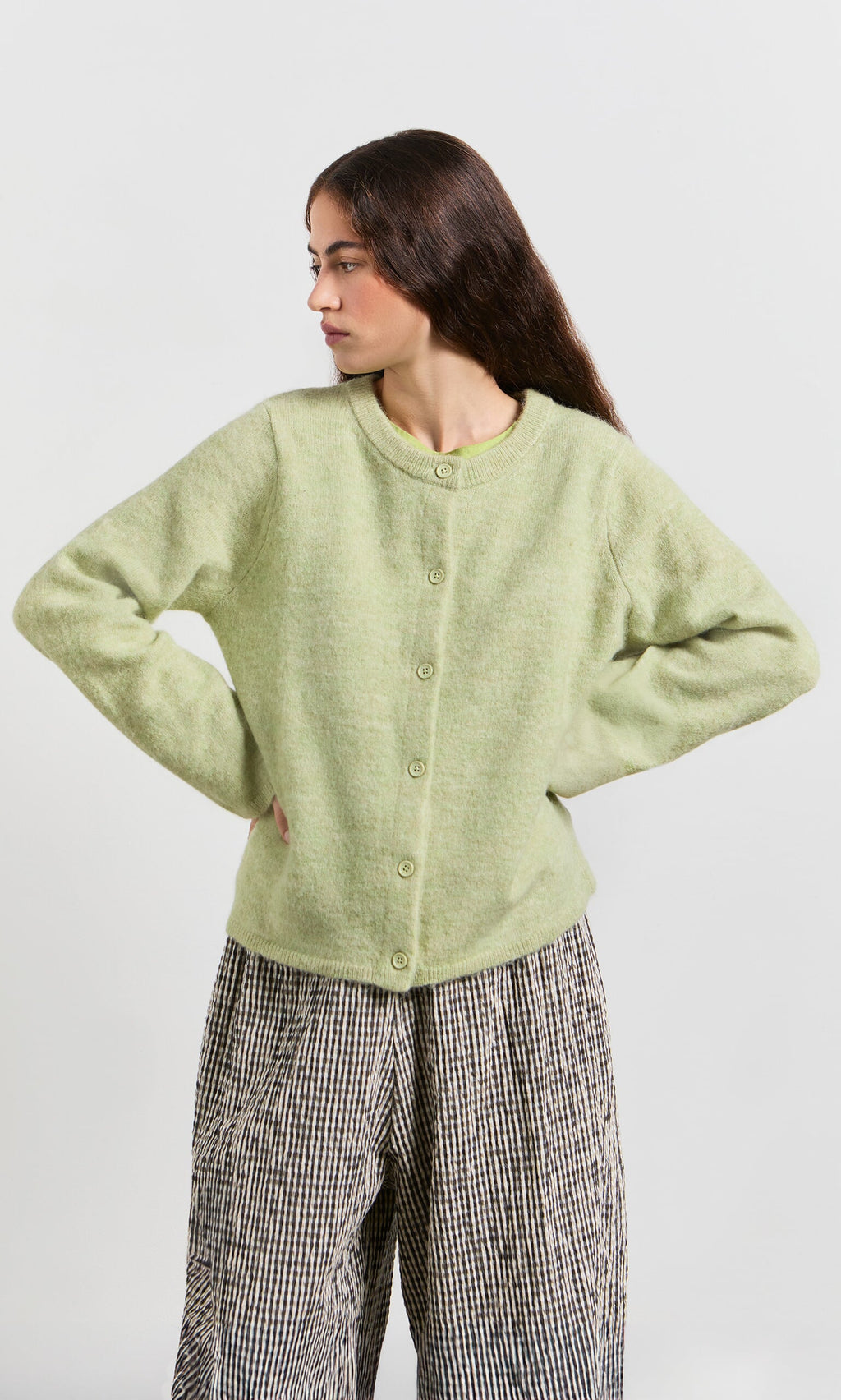 Plümo- Woman in a light green Pistaz Cardigan and loose checkered pants stands with hands on hips, looking to the side.
