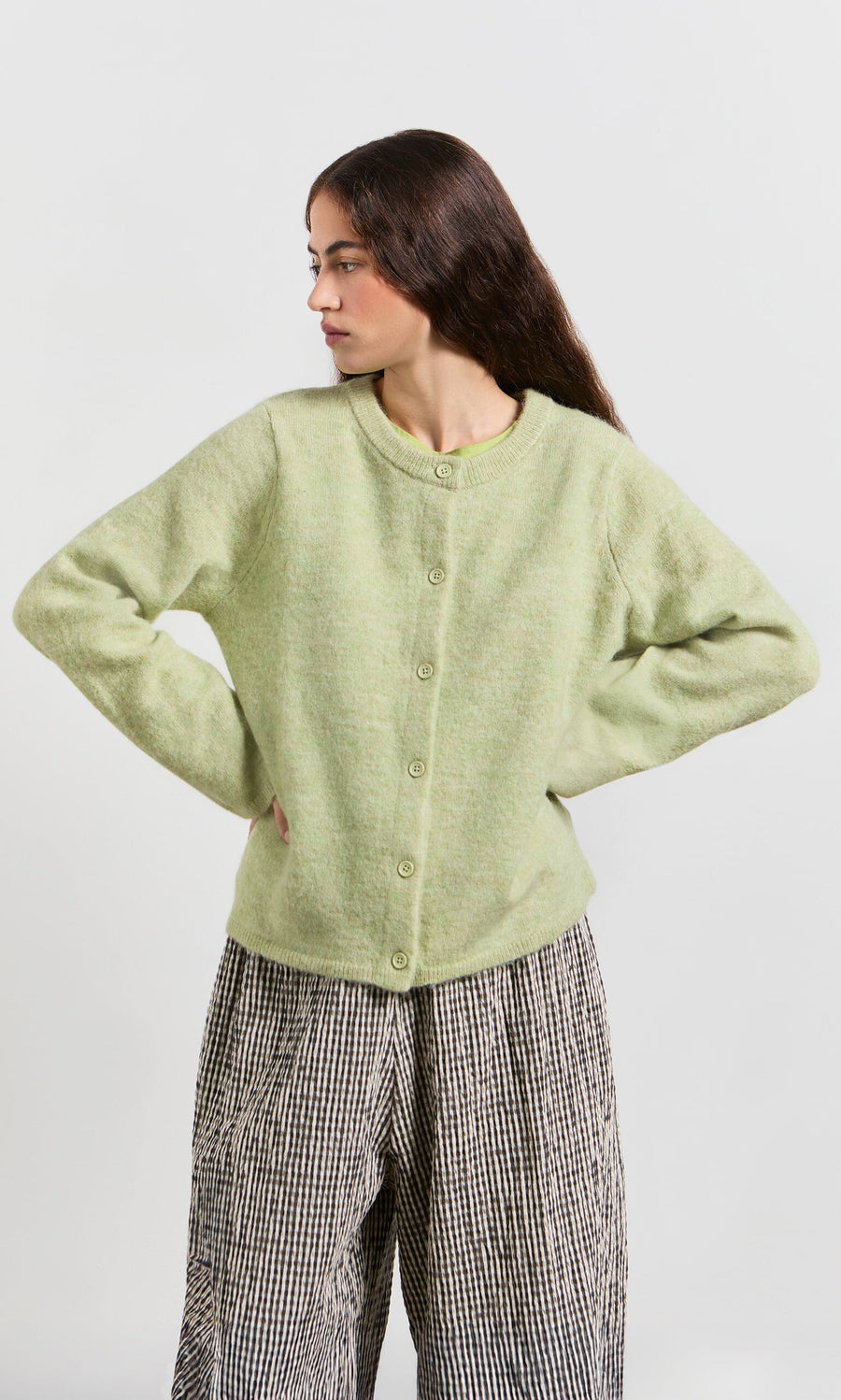 Plümo- Woman in a light green Pistaz Cardigan and loose checkered pants stands with hands on hips, looking to the side.