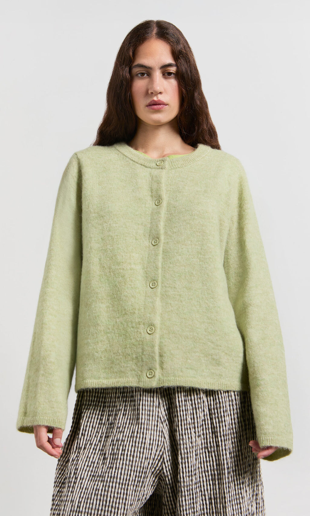 Plümo- Woman in a Pistaz Cardigan and checkered pants stands against a plain light background.