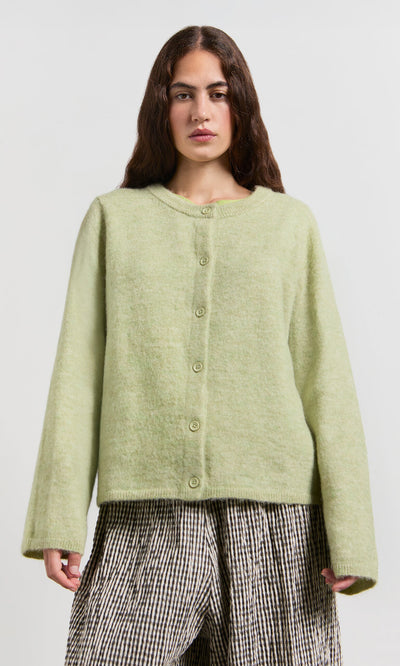Plümo- Woman in a Pistaz Cardigan and checkered pants stands against a plain light background.