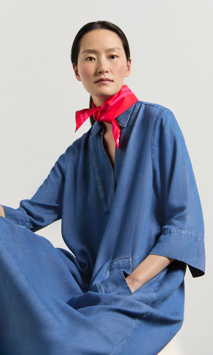 Plümo- A woman in the blue Portcullis Dress with pockets and a red scarf sits against a neutral background.