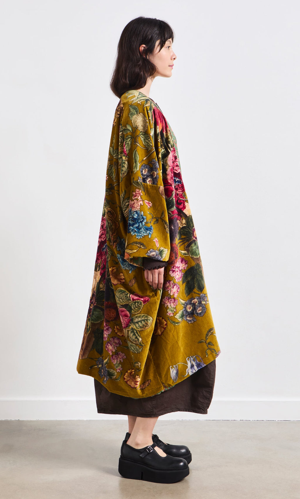 Plümo- Woman in a Pomfret Kimono, brown dress, and black platform shoes stands in profile against a white wall.