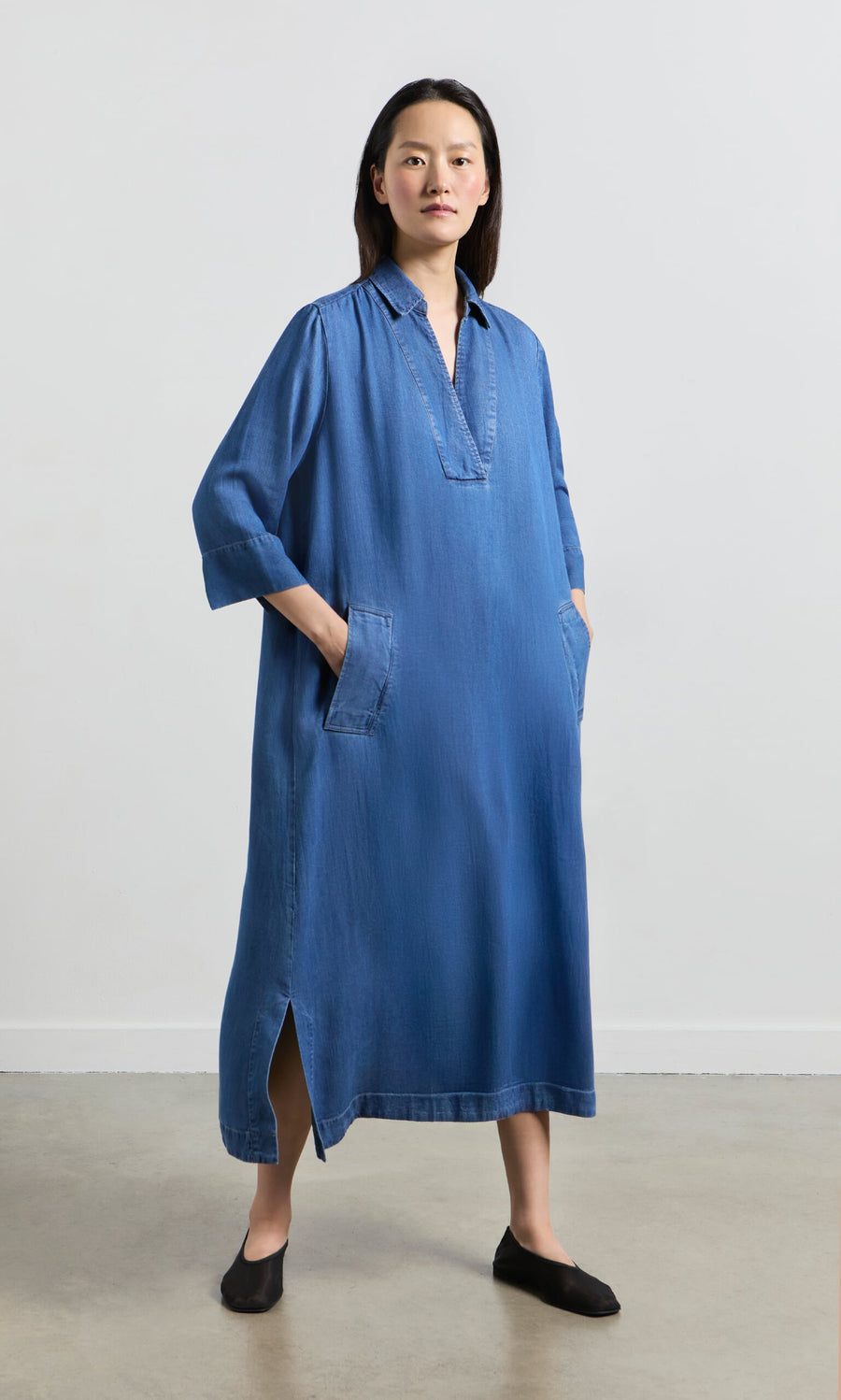 Plümo- Woman in a loose A-line blue Portcullis Dress with pockets, standing indoors on a light floor and background.