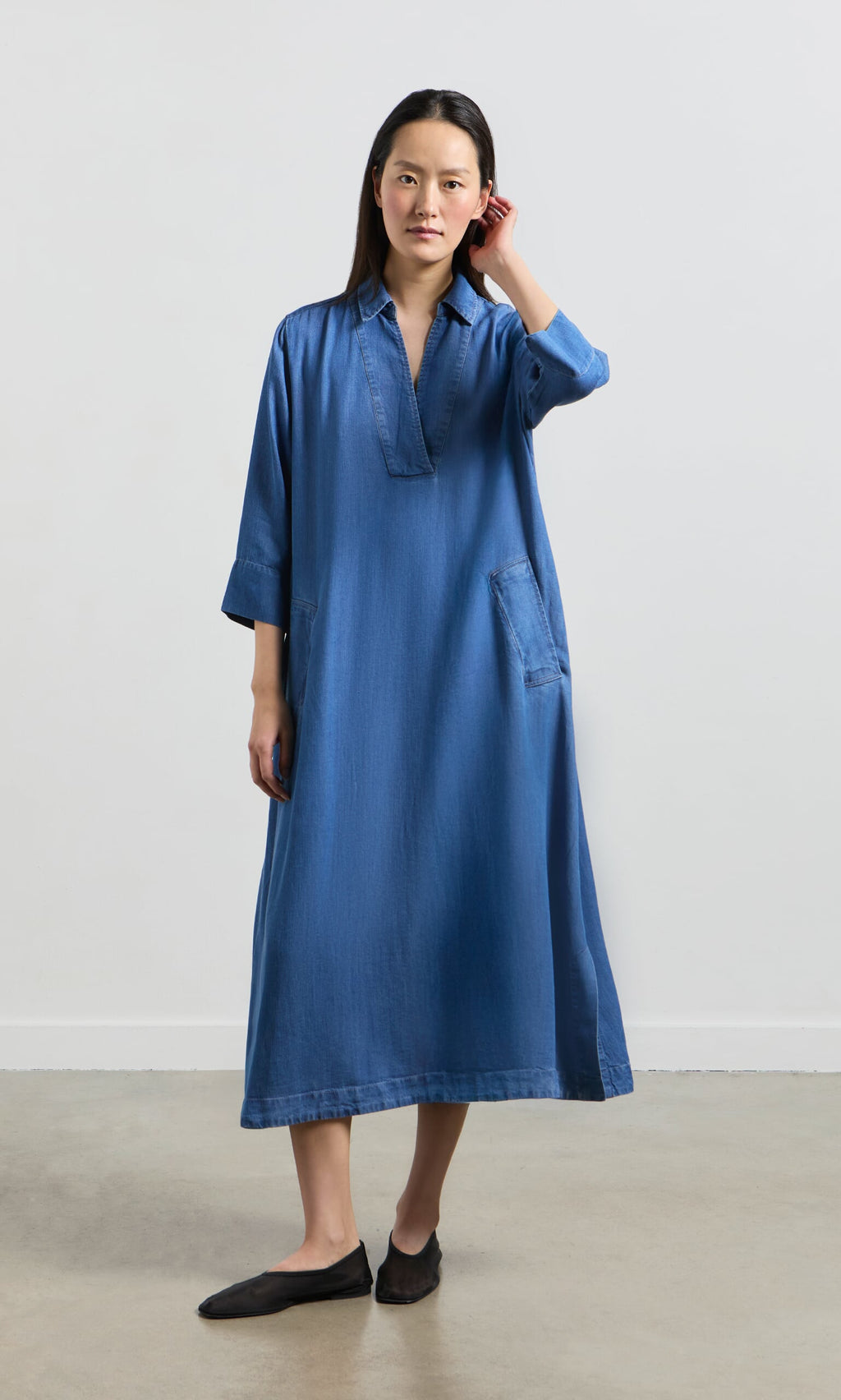 Plümo- Woman indoors wearing the Portcullis Dress, a loose A-line denim style with long sleeves and black flats, touching her hair.