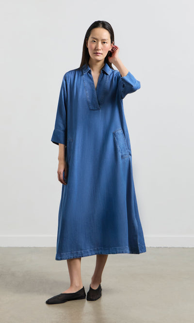 Plümo- Woman indoors wearing the Portcullis Dress, a loose A-line denim style with long sleeves and black flats, touching her hair.