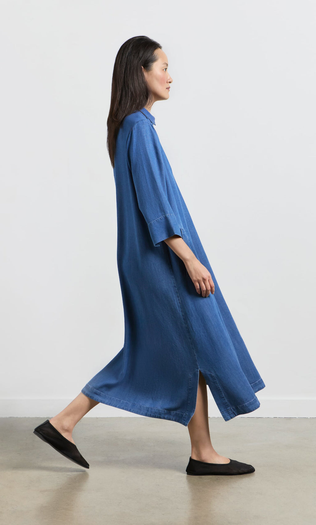 Plümo- Woman in the loose blue Portcullis Dress and black flats walks sideways on a light-colored floor.