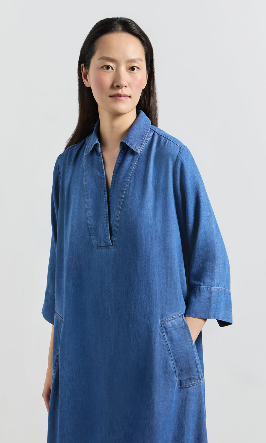 Plümo- Woman with long dark hair wears the Portcullis Dress, an A-line denim style, standing hands-in-pockets against a plain background.