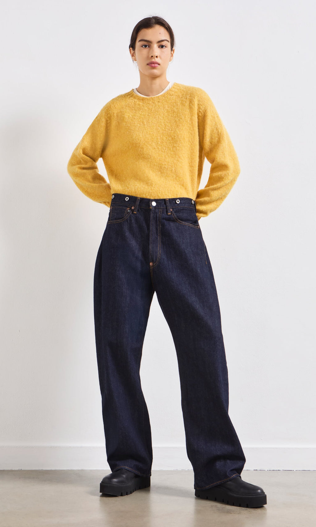 Plümo- A person in a yellow sweater, Japanese Raw Denim Jeans, and black shoes stands facing forward against a white wall.
