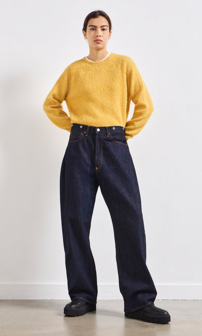 Plümo- A person in a yellow sweater, Japanese Raw Denim Jeans, and black shoes stands facing forward against a white wall.