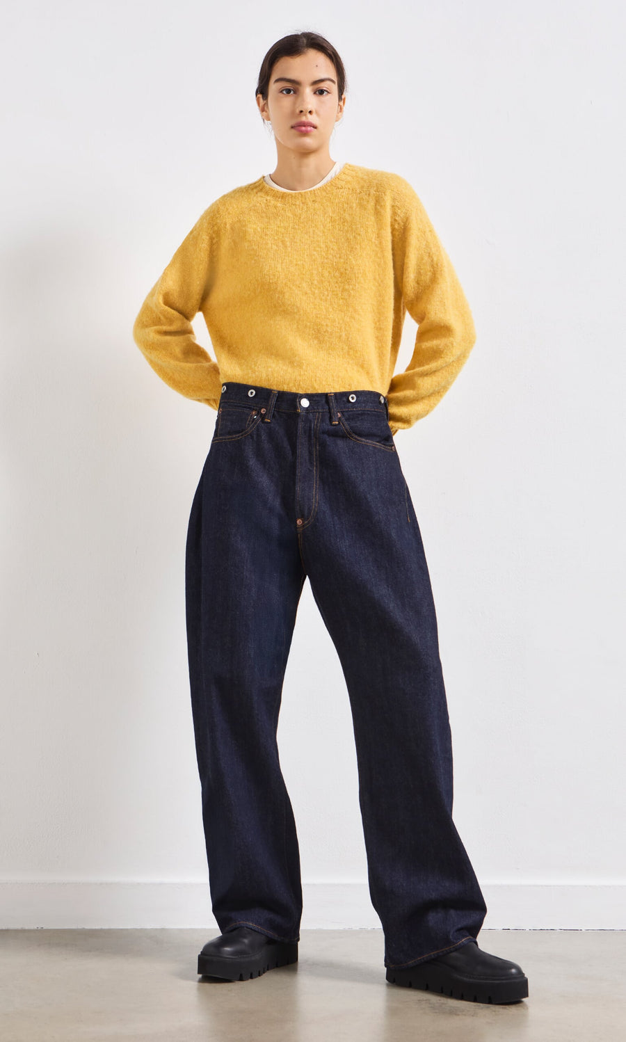 Plümo- A person in a yellow sweater, Japanese Raw Denim Jeans, and black shoes stands facing forward against a white wall.