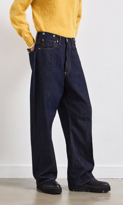 Plümo- Person in a yellow sweater, Japanese Raw Denim Jeans, and black shoes stands against a white wall.