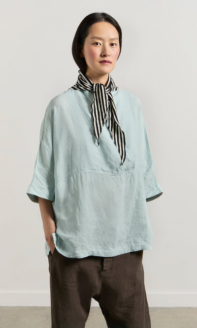 Plümo- A person wears a light blue top, brown pants, and the Lynn Stripe Necktie with hands in pockets.