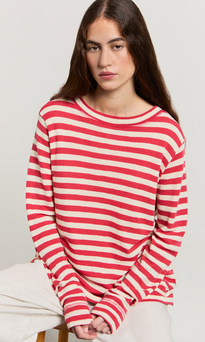 Plümo- Woman with long brown hair wears a Red Stripe Top and light pants, seated against a plain background.