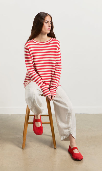Plümo- Woman in a Red Stripe Top, white pants, and red shoes sits on a wooden stool against a plain background.