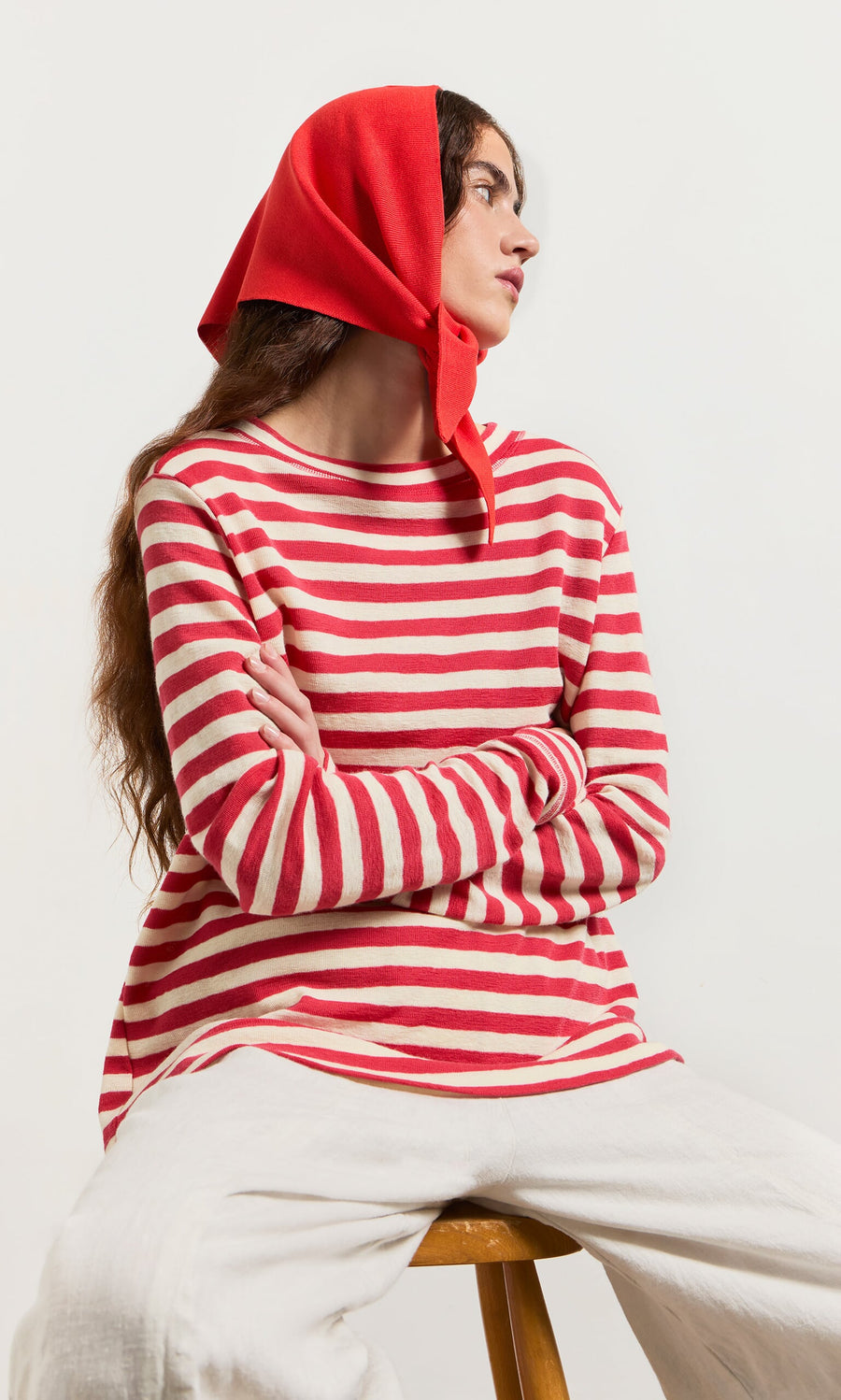 Plümo- Woman in a Red Stripe Top and red headscarf sits on a stool, looking to the side with arms crossed.