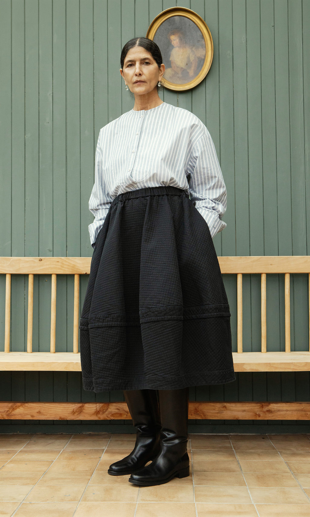 Plümo  Woman in a striped shirt and the Rho Skirt - Coal stands by a wooden bench, beneath a framed portrait on a green wall.
