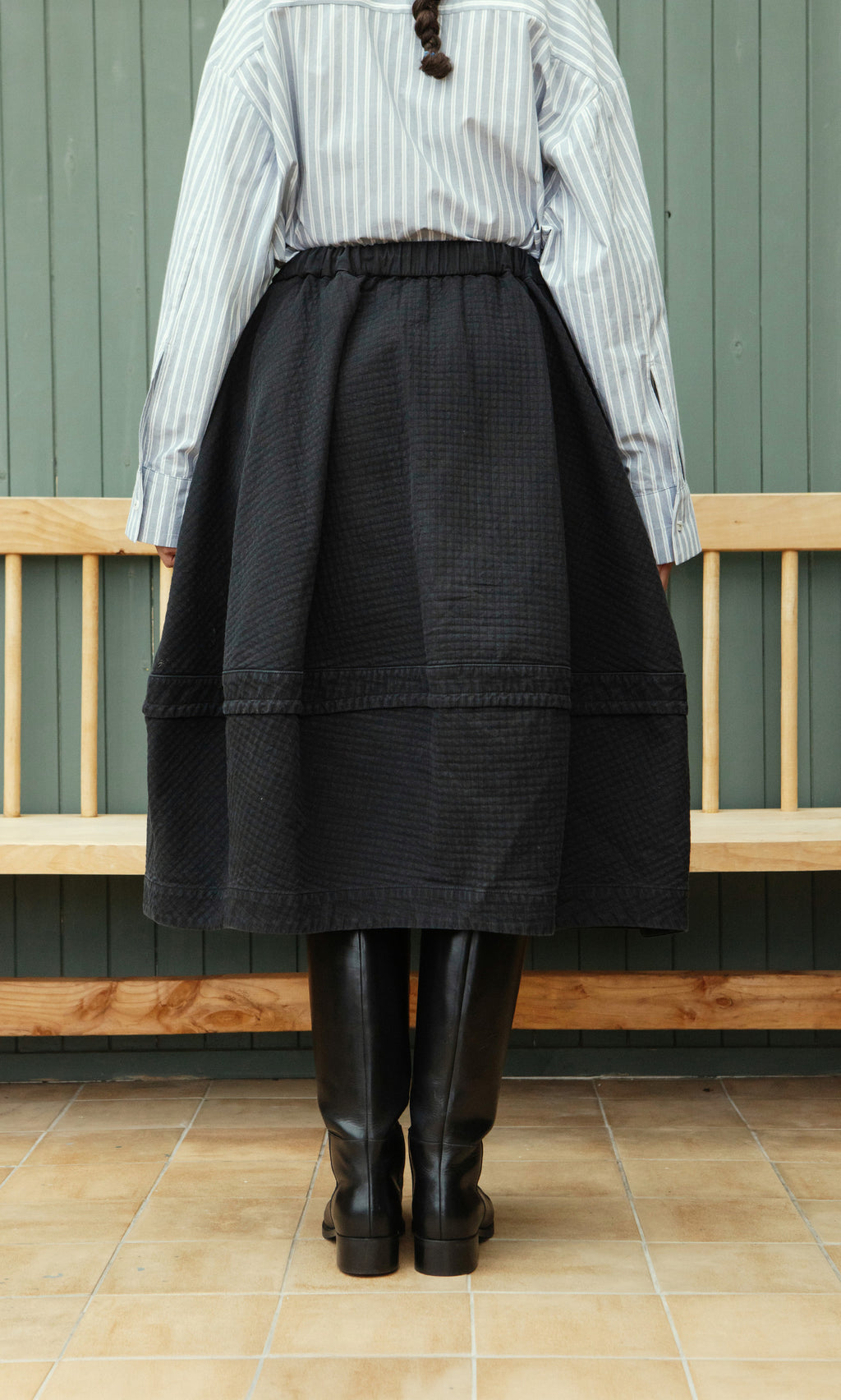 Plümo  Person in a striped shirt, Rho Skirt - Coal, and black boots stands before a wooden bench, facing away.