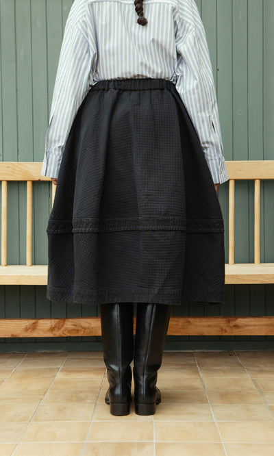 Plümo  Person in a striped shirt, Rho Skirt - Coal, and black boots stands before a wooden bench, facing away.