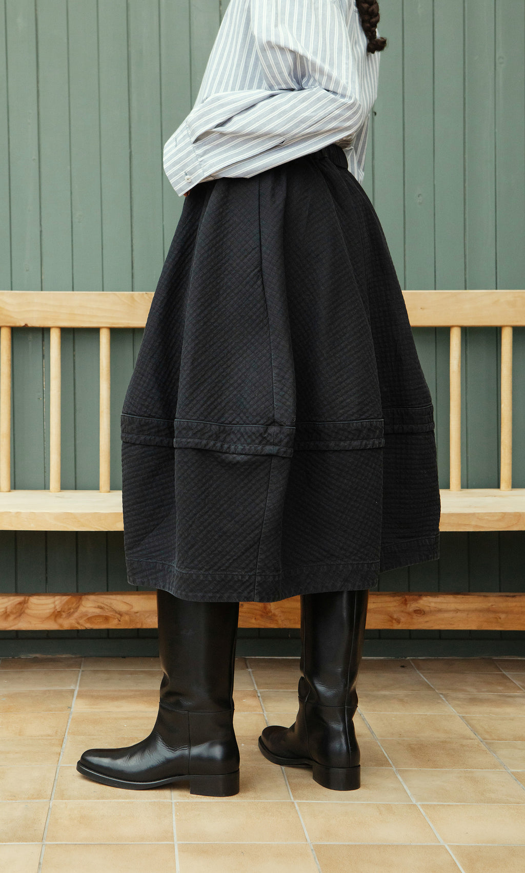 Plümo  Person in a striped shirt, Rho Skirt - Coal, and tall black boots stands sideways by a wooden bench indoors.