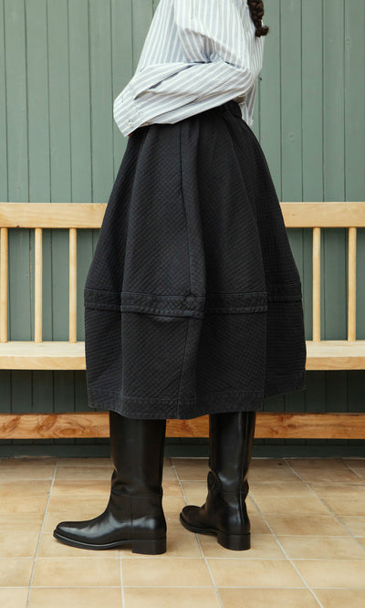 Plümo  Person in a striped shirt, Rho Skirt - Coal, and tall black boots stands sideways by a wooden bench indoors.