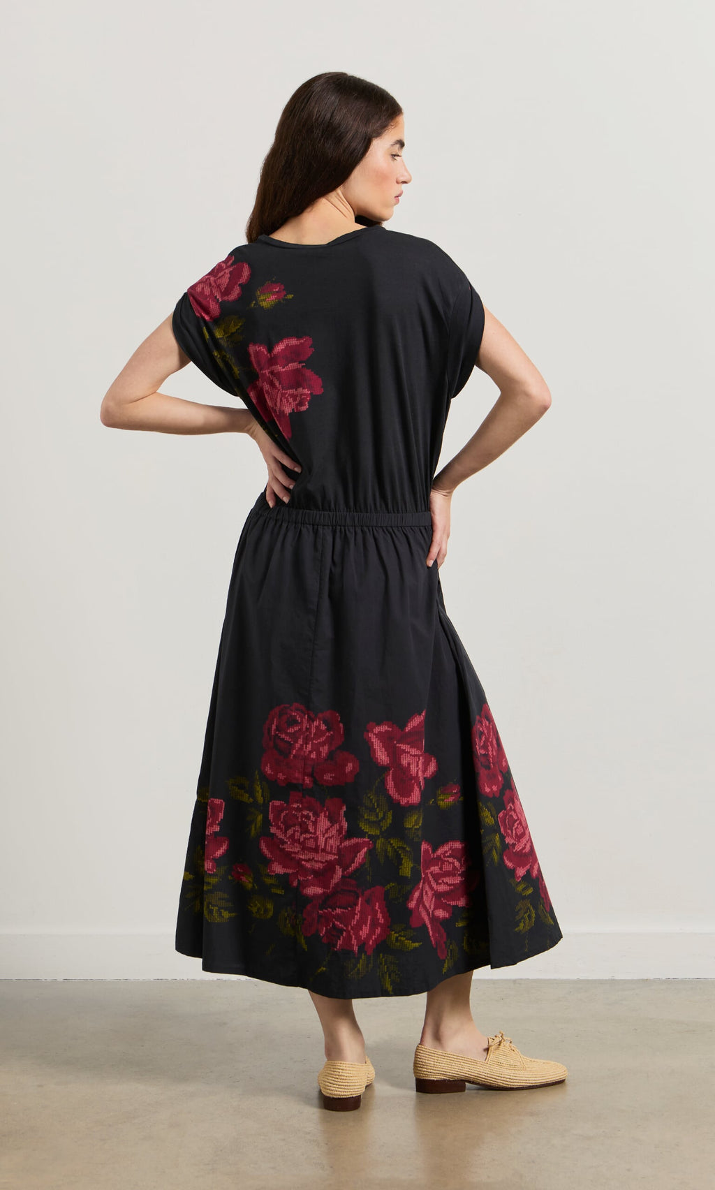 Plümo- Woman facing away in the Rose Stitch Dress, featuring black cotton with a large red rose design, and beige shoes.