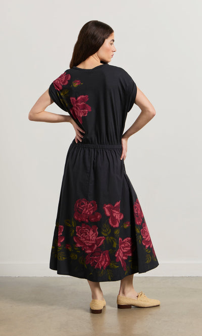 Plümo- Woman facing away in the Rose Stitch Dress, featuring black cotton with a large red rose design, and beige shoes.