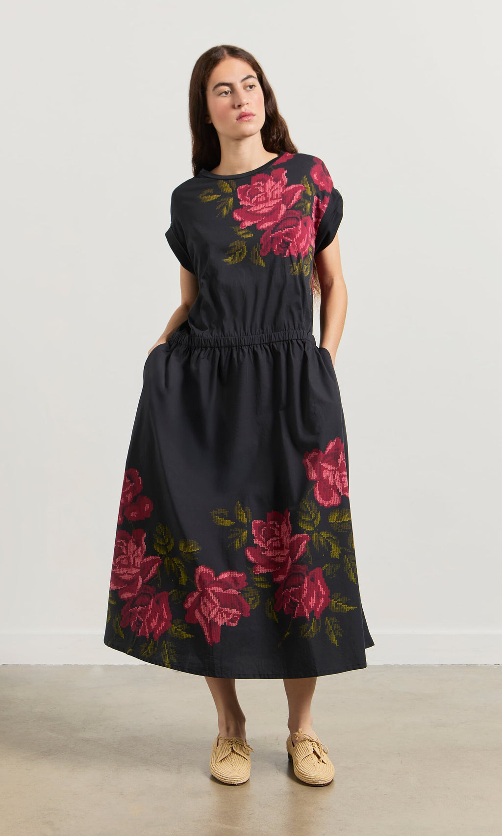 Plümo- Woman models the Rose Stitch Dress—black cotton with cross-stitch rose design—hands in pockets, beige shoes.