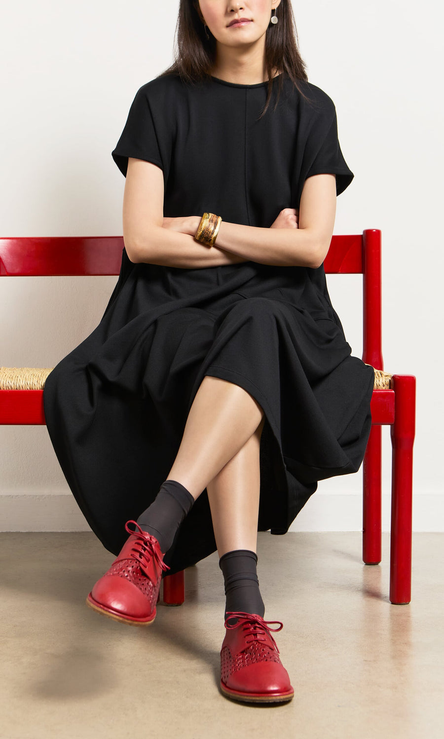 Plümo- Woman in a black dress, arms crossed, sits on a red bench wearing Magui Brogues and black socks.