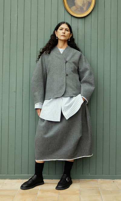 Plümo  Woman in grey Ghent Skirt and jacket, striped shirt visible, stands by green wall with framed art above.