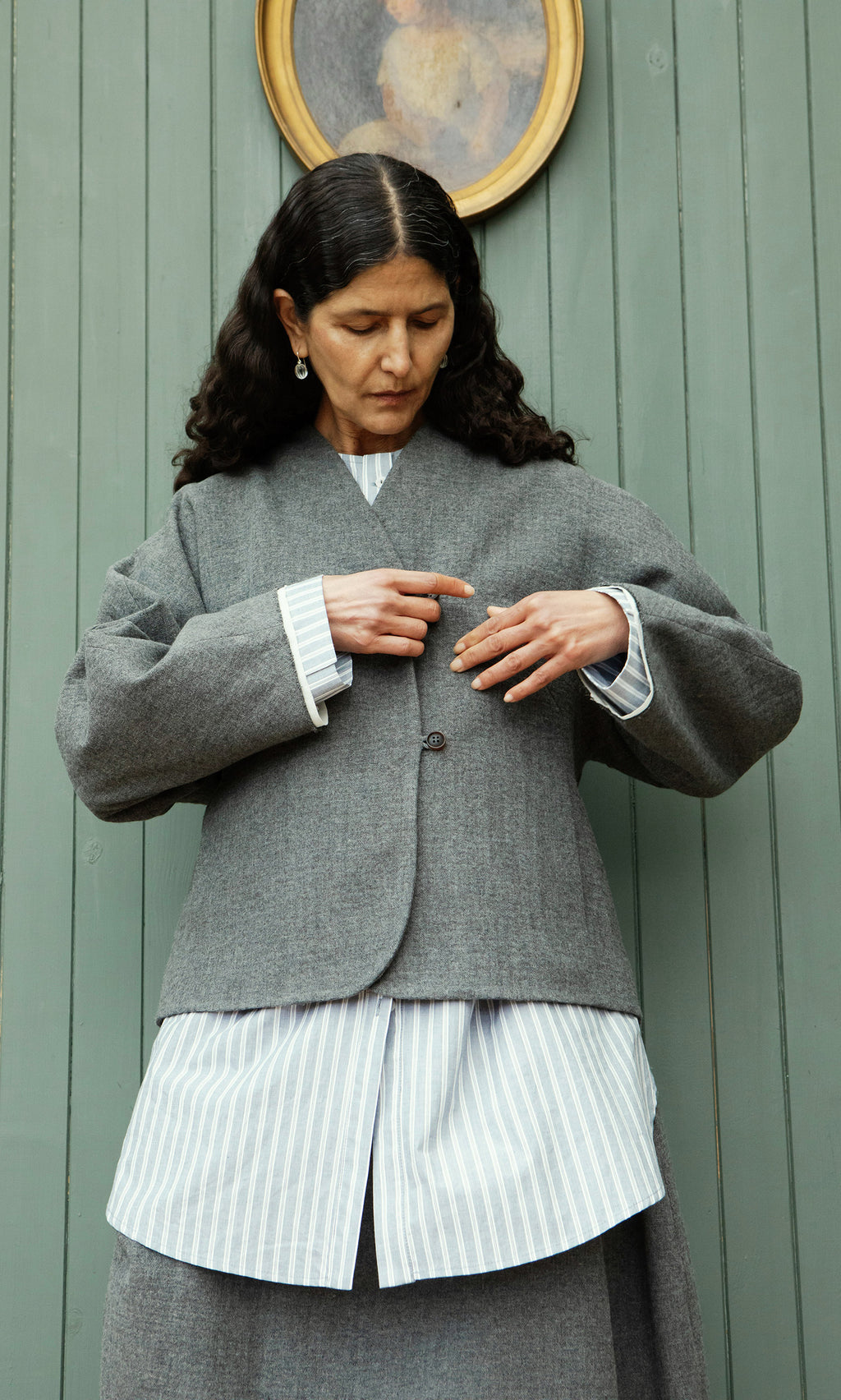 Plümo  Woman with long dark hair fastens a Rostock Jacket over a striped shirt, standing by a green wooden wall.
