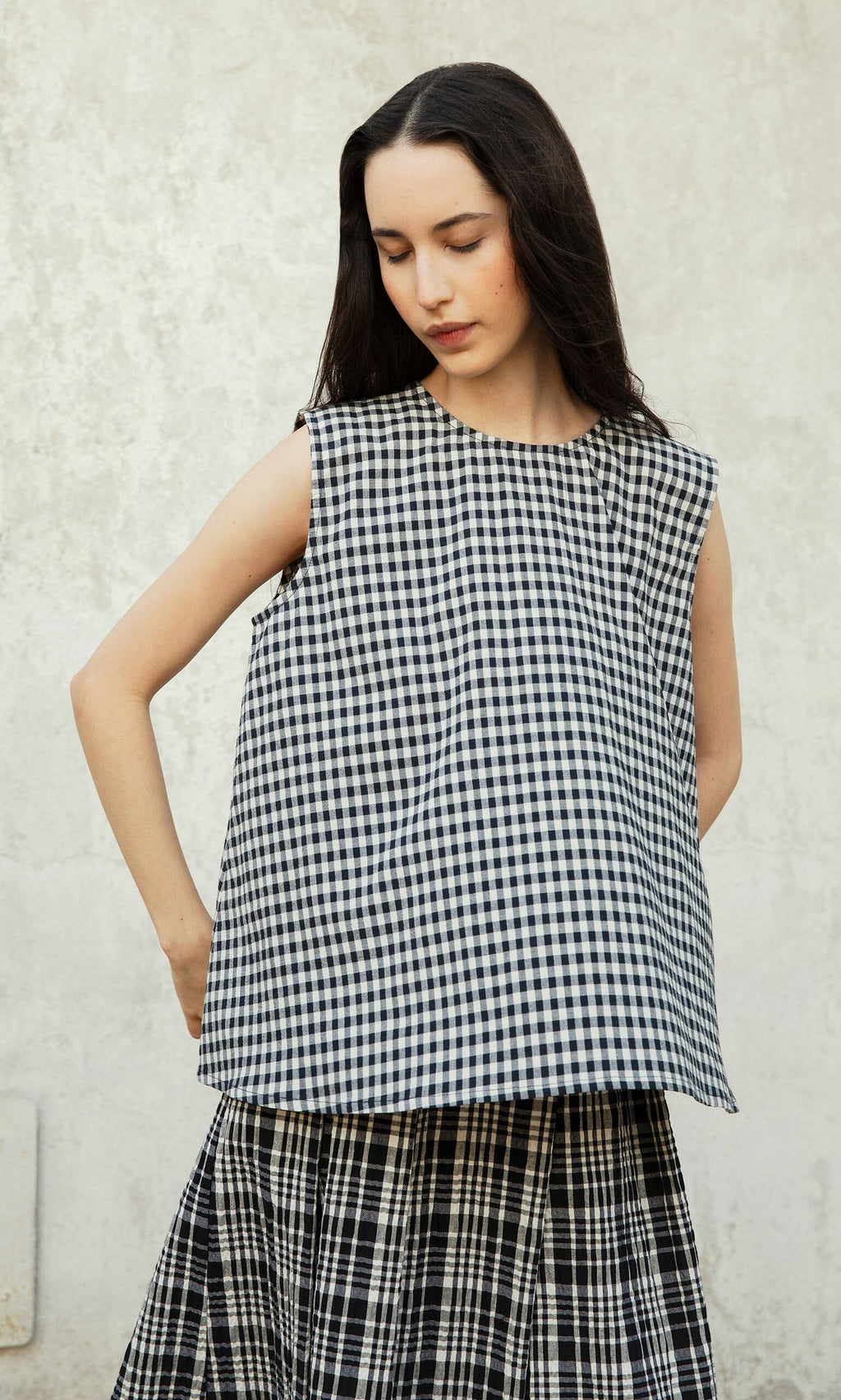 Plümo- Woman in Serena Top - Gingham with matching plaid skirt, standing against a light wall.