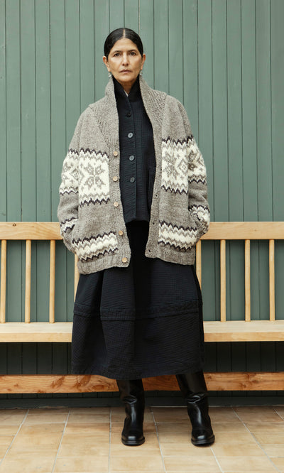 Plümo  Woman in a Shingle Cardigan over a black dress and boots stands by a bench and green paneled wall.