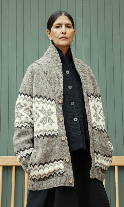 Plümo  A woman in the Shingle Cardigan with geometric patterns stands by a green wooden wall, looking serious.