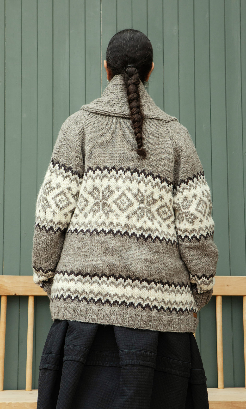 Plümo  Someone with a braid wears the Shingle Cardigan, standing back to the camera against a green wall.