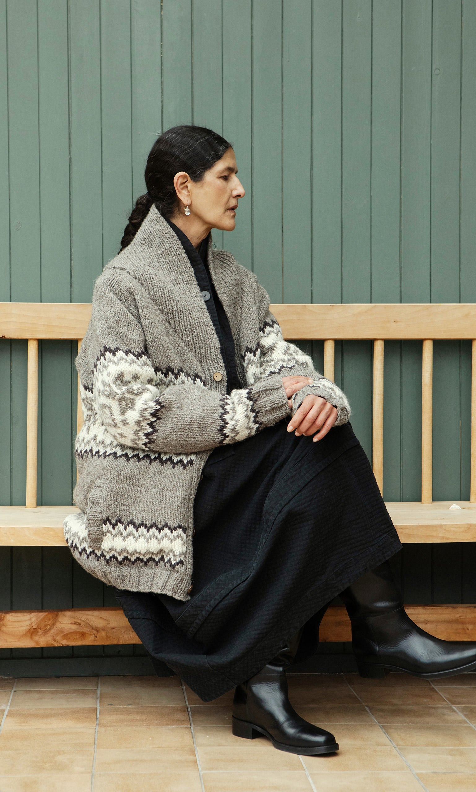 Plümo  Woman in a Shingle Cardigan and black dress sits sideways on a wooden bench against a green paneled wall.