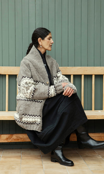 Plümo  Woman in a Shingle Cardigan and black dress sits sideways on a wooden bench against a green paneled wall.