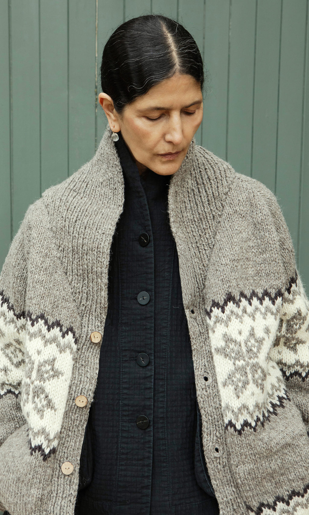 Plümo  A person with dark hair in a bun wears the Shingle Cardigan over a black shirt, looking down.