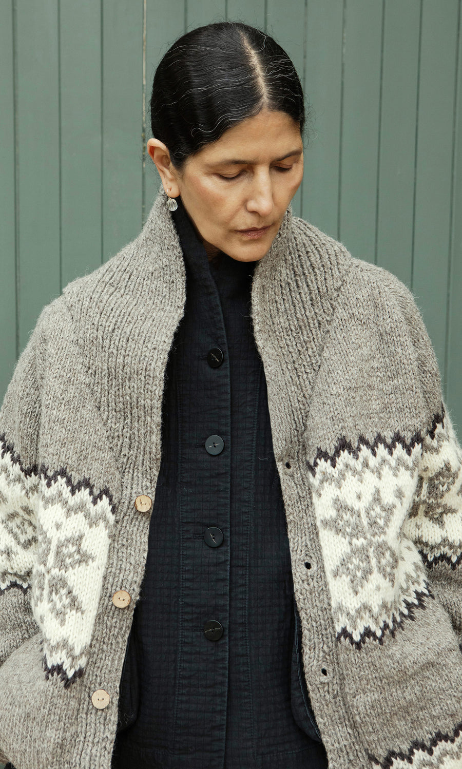 Plümo  A person with dark hair in a bun wears the Shingle Cardigan over a black shirt, looking down.