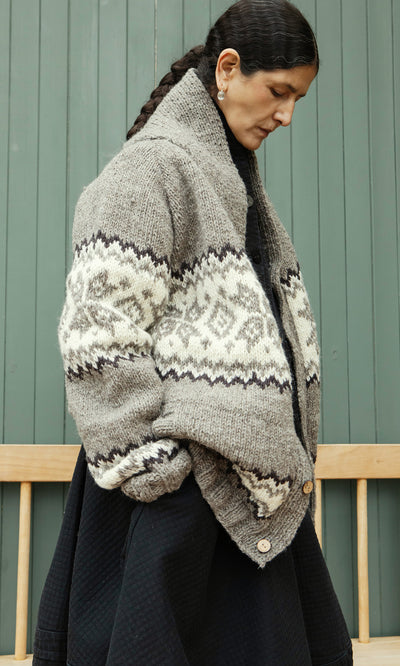 Plümo  Woman in a Shingle Cardigan stands sideways, looking down, before a green wooden wall.