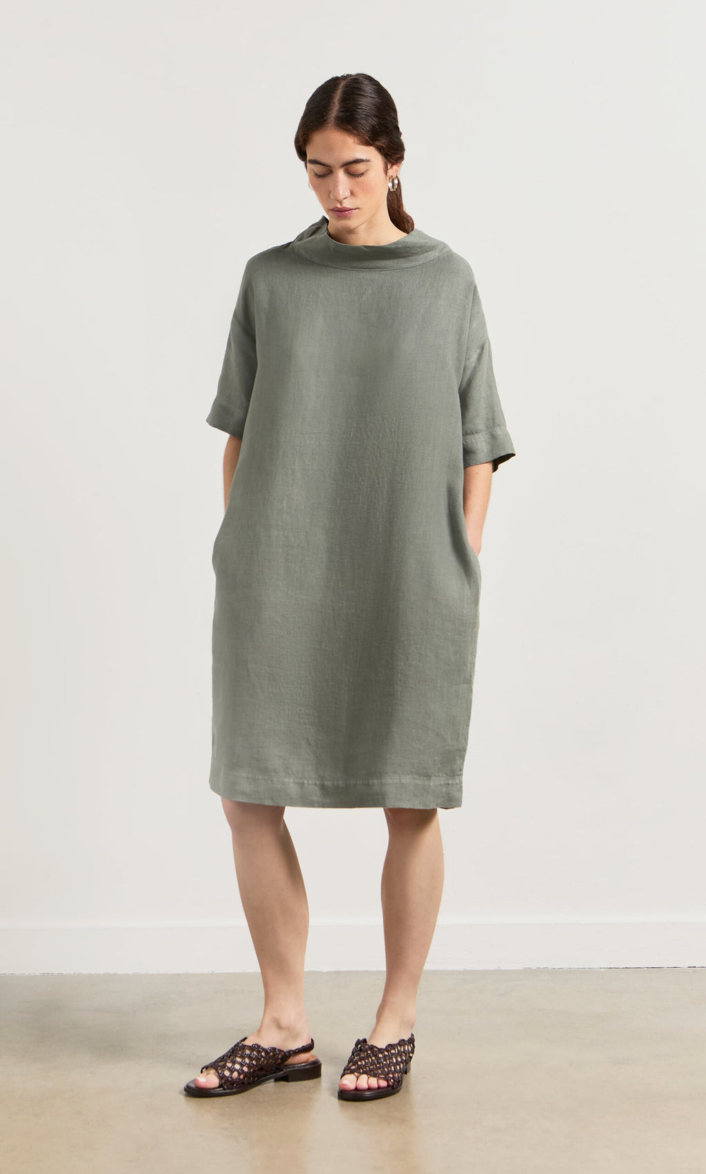 Plümo- Woman in the sage green Sims Dress—a loose linen shift with crop sleeves and pockets—pairing woven sandals.